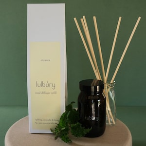 May include: A white box with the text "citraura" and "lulbúry reed diffuser refill" next to a dark brown glass bottle with a black lid and a small glass jar with wooden reeds. A sprig of green leaves is in front.