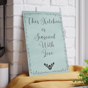 Peut inclure: Panneau de cuisine rectangulaire bleu clair avec l'inscription "This Kitchen is Seasoned With Love" en écriture cursive. Le panneau est orné d'une bordure florale noire et d'un petit motif floral en bas.