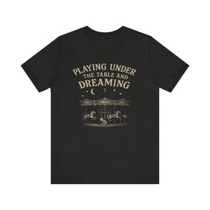 May include: Dark gray t-shirt with the text "PLAYING UNDER THE TABLE AND DREAMING" in a vintage font. The graphic features a carousel with horses and a crescent moon. A whimsical design for a casual tee.