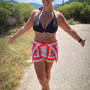 May include: Crochet shorts featuring a granny square pattern in neon orange, white, and grey yarn. The shorts have a drawstring waist, a high-cut leg design, and a visible texture created by the interlocking yarn stitches throughout the garment.