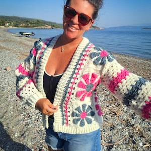 Puede incluir: Un cárdigan de crochet color crema con detalles florales rosas y grises. El suéter abierto tiene cuello en V y mangas largas. La mujer de la imagen lleva el cárdigan con pantalones cortos de mezclilla y gafas de sol en la playa.