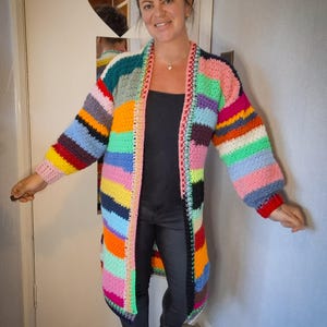 Op de afbeelding: Een lange, handgemaakte gehaakte cardigan in een patchwork-ontwerp. De cardigan heeft een levendige mix van kleuren, waaronder roze, groen, blauw, geel en oranje. De mouwen zijn lang en de cardigan komt tot halverwege de kuit. De afwerking is in een contrasterende kleur.