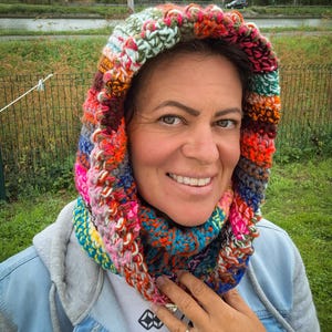 Op de afbeelding: Een veelkleurige, grof gehaakte capuchon en colset met een levendige mix van garenkleuren, waaronder oranje, blauw, roze, groen en wit. Het accessoire is gemaakt met een dik, gestructureerd steekpatroon dat het hoofd en de nek omlijst.