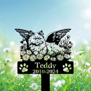 May include: Black metal pet memorial garden stake featuring a cat with angel wings, surrounded by flowers. The name "Teddy" and dates "2010-2024" are displayed below the cat, with paw prints on either side. The background is a blurred green and blue.