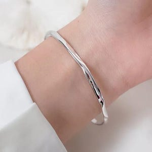 May include: A silver-coloured, textured cuff bracelet. The bracelet has a slightly irregular, organic shape, reflecting light. The jewellery is worn on a wrist, with a white shirt visible in the background. The bracelet is a simple, elegant accessory.