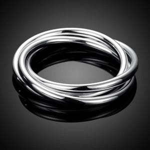 Può includere: Un braccialetto argentato con più fasce lisce e arrotondate, interconnesse. Le fasce sono riflettenti, creando riflessi e ombre. Il braccialetto è esposto su una superficie scura e lucida. Un gioiello.