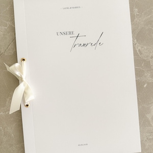 Könnte beinhalten: Ein weißes Hochzeitsprogramm mit dem Text "UNSERE Traurede" in eleganter Schrift. Der Umschlag ist mit einem cremefarbenen Band gebunden und mit goldenen Ösen befestigt. Die Namen "LAURA & MARKUS" und das Datum "16.06.2026" sind ebenfalls aufgedruckt.