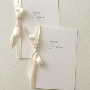 Può includere: Due biglietti bianchi con nastri di raso color crema annodati. I biglietti presentano le frasi "AN MEINE Ehefrau" e "AN MEINEN Ehemann" in grafia nera. I biglietti sono probabilmente per un matrimonio o un anniversario.