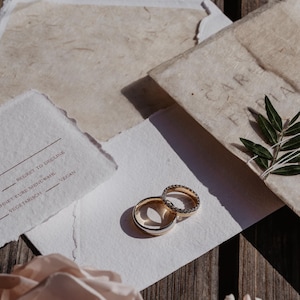 Pu&ograve; includere: Due fedi nuziali, una con una fascia di diamanti, sono appoggiate su un biglietto bianco con testo. Il biglietto &egrave; accanto a un fiore rosa chiaro e cancelleria. Lo sfondo &egrave; una superficie di legno.