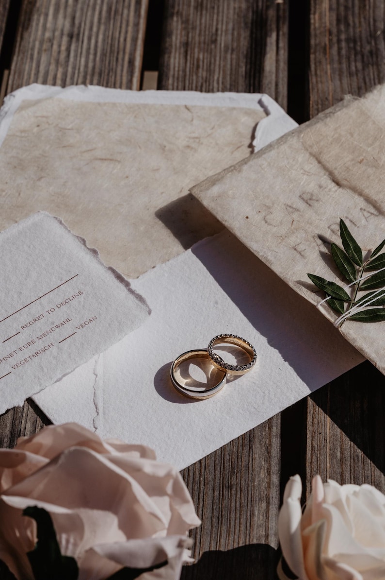 Pu&ograve; includere: Due fedi nuziali, una con una fascia di diamanti, sono appoggiate su un biglietto bianco con testo. Il biglietto &egrave; accanto a un fiore rosa chiaro e cancelleria. Lo sfondo &egrave; una superficie di legno.