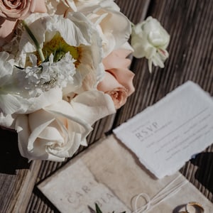 Pu&ograve; includere: Una suite di inviti di nozze con un bouquet di rose e peonie bianche e rosa. La suite comprende una scheda RSVP, un invito avvolto con il testo "CAR FABIA" e due fedi nuziali d'oro. Gli articoli sono disposti su una superficie di legno.