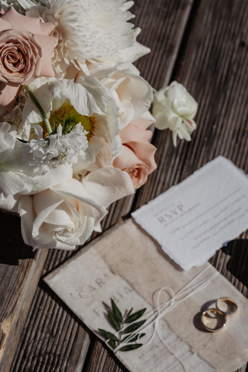 Pu&ograve; includere: Una suite di inviti di nozze con un bouquet di rose e peonie bianche e rosa. La suite comprende una scheda RSVP, un invito avvolto con il testo "CAR FABIA" e due fedi nuziali d'oro. Gli articoli sono disposti su una superficie di legno.