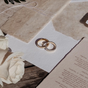 Pu&ograve; includere: Due fedi nuziali d'oro, una con una fascia di piccoli diamanti, poggiano su un biglietto bianco. Gli anelli sono accanto a un invito di nozze con testo e un rametto di verde. Una rosa bianca &egrave; nell'angolo.