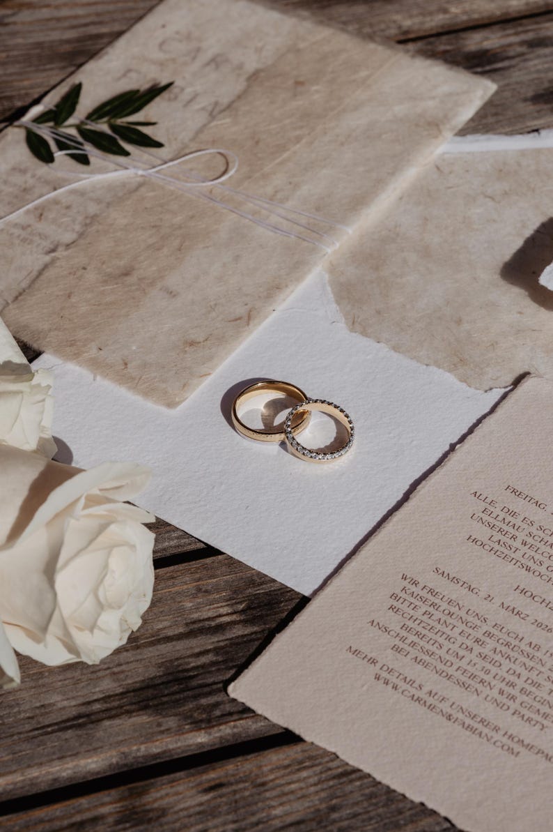Pu&ograve; includere: Due fedi nuziali d'oro, una con una fascia di piccoli diamanti, poggiano su un biglietto bianco. Gli anelli sono accanto a un invito di nozze con testo e un rametto di verde. Una rosa bianca &egrave; nell'angolo.