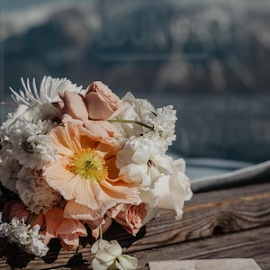 Pu&ograve; includere: Un bouquet da sposa con papaveri color pesca, rose rosa e fiori bianchi &egrave; appoggiato su una superficie di legno. Fedi nuziali e un biglietto RSVP suggeriscono un tema nuziale.