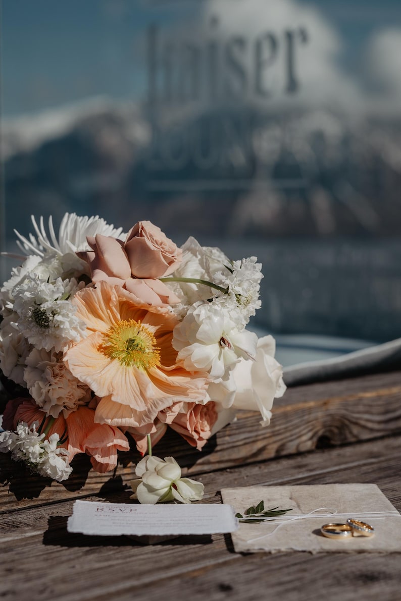 Pu&ograve; includere: Un bouquet da sposa con papaveri color pesca, rose rosa e fiori bianchi &egrave; appoggiato su una superficie di legno. Fedi nuziali e un biglietto RSVP suggeriscono un tema nuziale.