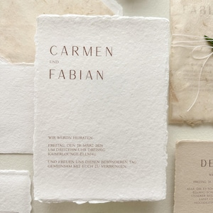 Pu&ograve; includere: Un insieme di inviti di nozze in carta fatta a mano. L'invito principale presenta i nomi "CARMEN UND FABIAN" in marrone. Sono inclusi data, ora e luogo del matrimonio. La carta ha un aspetto strutturato e bianco sporco.