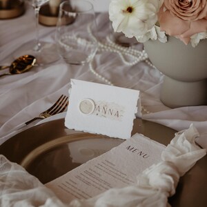 Pu&ograve; includere: Elegante mise en place con un piatto dorato, lino bianco e una composizione floreale di rose bianche e rosa. &Egrave; esposto un segnaposto con il nome "ANNA" e un menu.