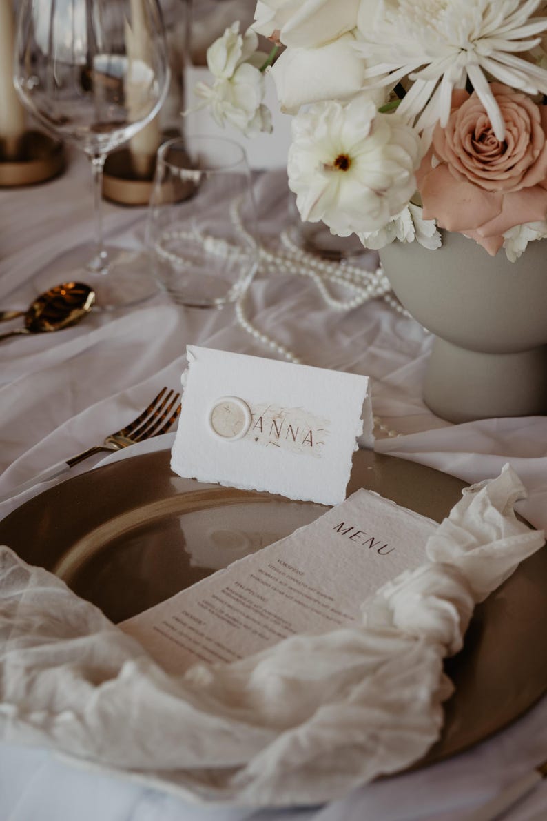 Pu&ograve; includere: Elegante mise en place con un piatto dorato, lino bianco e una composizione floreale di rose bianche e rosa. &Egrave; esposto un segnaposto con il nome "ANNA" e un menu.