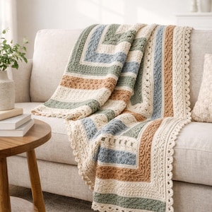 May include: A handcrafted, multi-colored crochet blanket draped over a beige sofa. The blanket features stripes of cream, blue, green, and brown, with a textured stitch pattern and a scalloped edge. A small wooden side table and a vase with greenery are in the foreground.