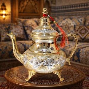 Könnte beinhalten: Eine goldfarbene Teekanne mit aufwendigen silbernen Details und einer roten Quaste. Die Teekanne hat einen gebogenen Ausguss, einen Griff und steht auf drei kleinen Füßen. Das Design ist wahrscheinlich von nahöstlichen oder nordafrikanischen Kulturen inspiriert.