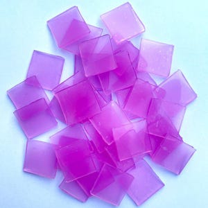 May include: A pile of translucent, square-shaped, purple glass tiles. The tiles are scattered on a light blue surface, with varying shades of purple creating depth. These tiles are suitable for mosaic art projects.