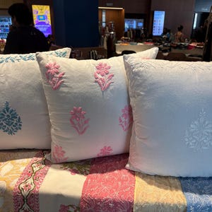 Könnte beinhalten: Drei weiße Zierkissen mit gestickten Blumenmustern in Rosa und Blau. Die Kissen sind auf einer farbenfrohen Patchwork-Oberfläche angeordnet und zeigen eine Vielzahl von Farben und Mustern.