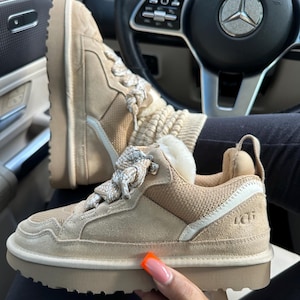 May include: Tan suede sneakers with a thick, off-white sole. The sneakers feature a mesh and suede upper, white laces, and the brand name "UGG" embossed on the side. The shoes are shown inside a car.