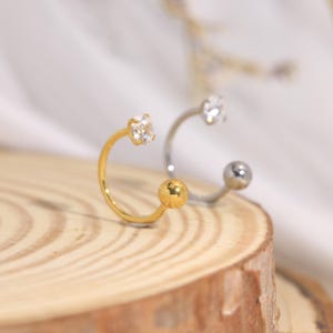 May include: Two open hoop earrings, one gold-toned with a clear faceted stone and one silver-toned with a smooth spherical end. Each earring features a circular band design with a small decorative element at one end and a larger terminal piece.