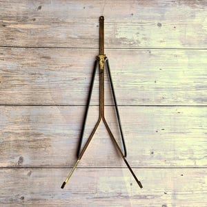 May include: A vintage brass and black easel with a tripod base. The easel has a decorative top and is designed to hold a small painting or artwork. The legs are black and the frame is brass-colored.