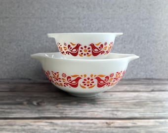 May include: Two white Pyrex mixing bowls stacked on top of each other. The bowls have a decorative pattern of red and orange birds, flowers, and leaves. The bowls have handles.