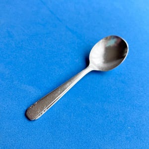 May include: A silver spoon with a rounded bowl and a long handle, resting on a bright blue surface. The handle has a decorative design near the end. The spoon is likely made of stainless steel.