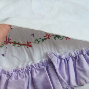 May include: Close-up of a white fabric with embroidered floral details in red, green, and purple. A lavender ruffled trim is visible at the bottom, adding a delicate touch to the textile.