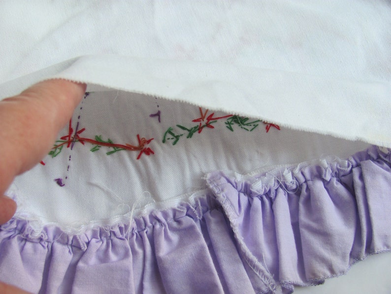 May include: Close-up of a white fabric with embroidered floral details in red, green, and purple. A lavender ruffled trim is visible at the bottom, adding a delicate touch to the textile.