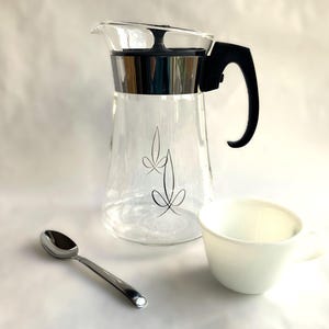 May include: A clear glass coffee carafe with a black handle and lid, a white cup, and a silver spoon. The carafe has a decorative black floral design. The items are arranged on a white surface.