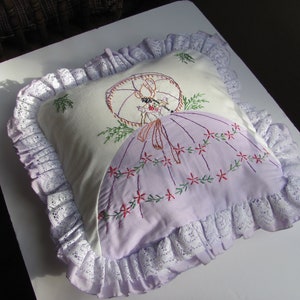 May include: A decorative pillow with a white background and a lavender skirt design. The pillow features an embroidered image of a woman with a parasol. The pillow is trimmed with white lace.