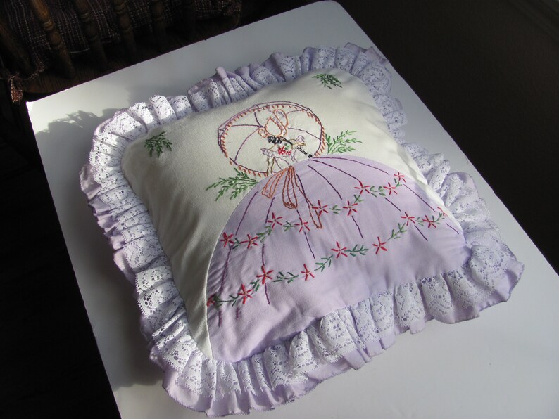 May include: A decorative pillow with a white background and a lavender skirt design. The pillow features an embroidered image of a woman with a parasol. The pillow is trimmed with white lace.