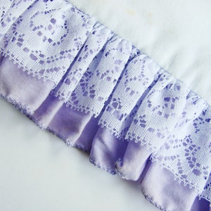 May include: Close-up of a fabric detail featuring a ruffled edge. The edge is composed of a lavender fabric base with a white lace overlay. The lace has a detailed floral pattern. The background is a light, off-white fabric.