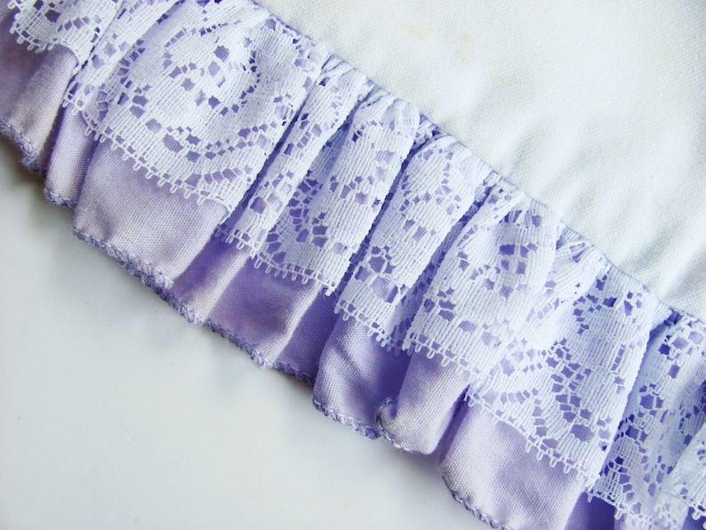 May include: Close-up of a fabric detail featuring a ruffled edge. The edge is composed of a lavender fabric base with a white lace overlay. The lace has a detailed floral pattern. The background is a light, off-white fabric.
