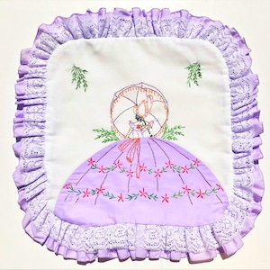 May include: Square white embroidered doily with a lavender ruffled lace edge. The center features a woman with an umbrella, wearing a lavender dress with red and green floral accents. The design is hand-stitched.