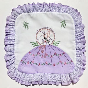 May include: Square white and lavender embroidered pillow cover with a vintage design. The pillow features a lavender dress with red floral accents, a woman with an umbrella, and a ruffled lace border. The pillow is approximately 12 inches square.