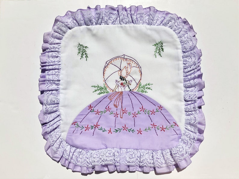 May include: Square white and lavender embroidered pillow cover with a vintage design. The pillow features a lavender dress with red floral accents, a woman with an umbrella, and a ruffled lace border. The pillow is approximately 12 inches square.