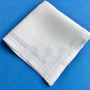 May include: A folded white linen handkerchief with a decorative embroidered border. The handkerchief is square and lies on a blue surface. The embroidered border features a repeating geometric pattern.