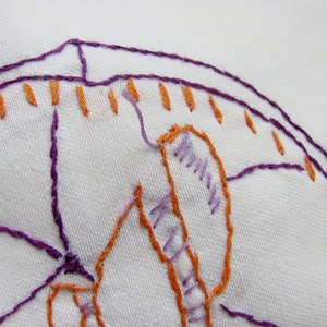 May include: Close-up of white fabric with embroidered design. The embroidery features purple and orange thread, creating a detailed pattern. The stitches are visible, showcasing the craftsmanship of the textile art.