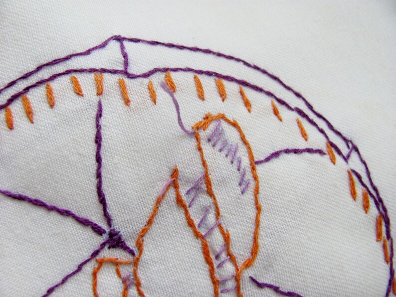 May include: Close-up of white fabric with embroidered design. The embroidery features purple and orange thread, creating a detailed pattern. The stitches are visible, showcasing the craftsmanship of the textile art.