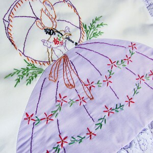 May include: Embroidered white and lavender fabric with a parasol design. The lavender parasol is decorated with red flowers and green vines. The embroidery features a figure with an umbrella and floral accents. The edges are trimmed with white lace.