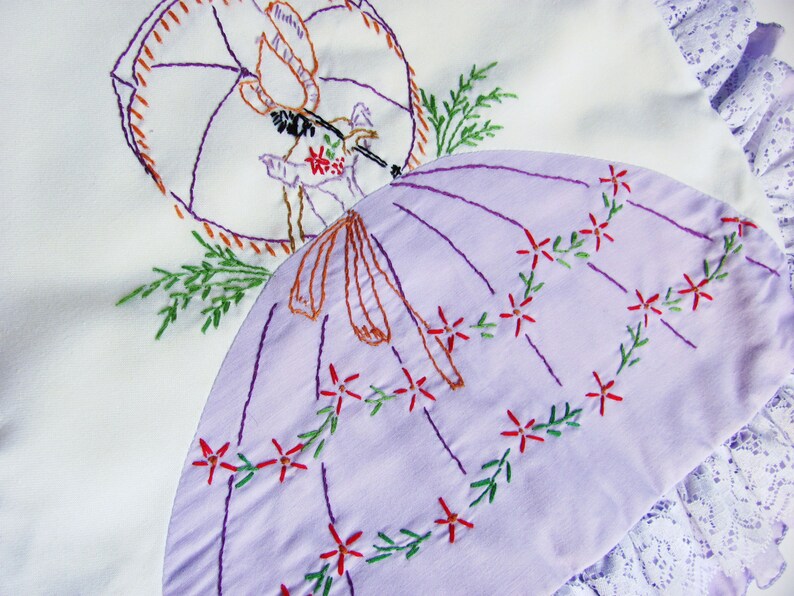 May include: Embroidered white and lavender fabric with a parasol design. The lavender parasol is decorated with red flowers and green vines. The embroidery features a figure with an umbrella and floral accents. The edges are trimmed with white lace.