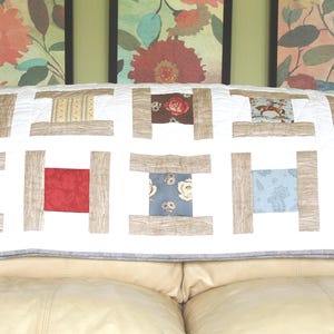 May include: A white quilted throw with a brown and beige geometric pattern. The throw is on a beige couch.