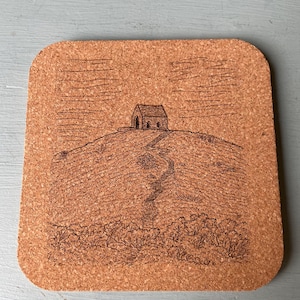 May include: Square cork coaster with a detailed etched design. The image depicts a small building atop a hill, with a path leading up to it. The coaster has a natural brown color and a slightly textured surface.