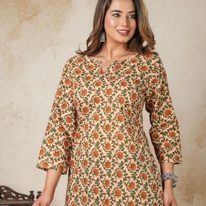Puede incluir: Top beige con estampado floral en naranja y verde. El top tiene cuello en V y mangas tres cuartos. La modelo lleva vaqueros azules. El top es de estilo casual.